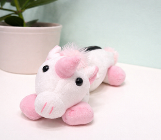 A white plush unicorn with pink hooves and horn rests on a tabletop; a simple plant pot and a soft green wall create a calm background.