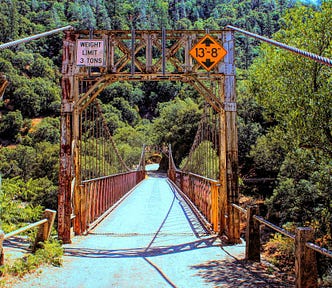 Old rusty single-lane gravel-covered suspension bridge with weight limit limit sign reading maximum of three-tons and an orange warning sign noting maximum height of thirteen feet, eight inches. The image was taken straight on down the center of the roadway with the thick bradded wires projecting out from the bridge to out of sight past the photographer. The bridge road empties onto a single road into a steep hillside thickly covered by trees and brush. The true length distance is deceiving.