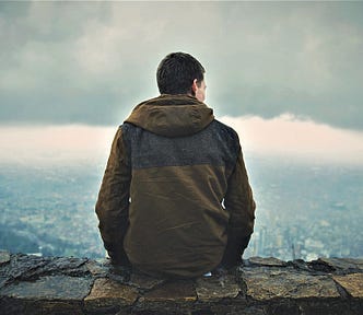 rear view of man wearing brown hooded sweatshirt sitting on stone wall looking out over valley