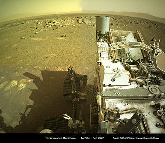 The Perseverance Rover on Mars takes a picture of the Martian landscape. The Rover takes up most of the right side of the image, showing off its intricate parts and instruments. Hills and maybe mountains can be seen in the distance as well as a variety of rocks of different colors, shapes and sizes littering the Martian plain it is in.