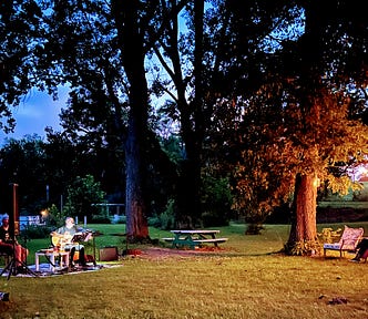 Two acoustical musicians, one on guitar and the other playing harmonica performing lakeside under an evening sky.