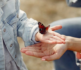 Photograph of a young girl holding a butterfly in her outstretched hand. The child’s mother is cupping her hand below the child’s.
