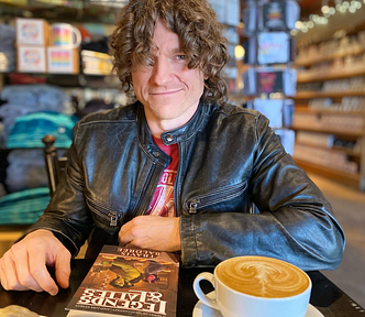 Travis Baldree, middle-aged, white, curly brunette hair, sits with his book and a latte.