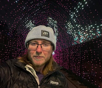 The author is surrounded by white and purple lights. He’s wearing a knitted hat that says “Joy as resistance.” He’s wearing black rimmed glasses and has a beard with enough white to betray that he’s middle-aged. He has long brown hair and a black coat that says “Denver” over a purple sweater. Purple glitter eyeliner matches the lights behind him.