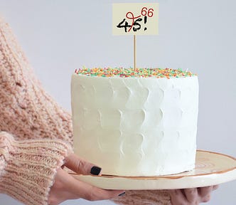 A woman’s arms holding a white birthday cake with a sign that says 45!, crossed out and corrected to 66.