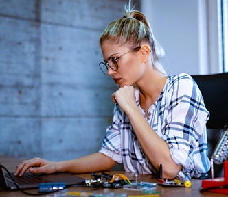 Woman with blonde ponytail and glasses sitting at a desk working on a laptop