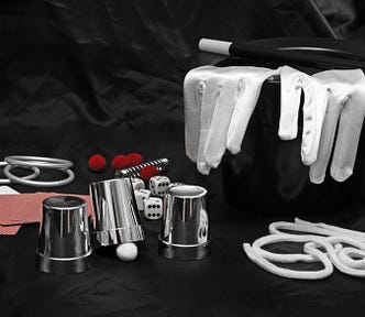 Color photo of the magician’s tools of the trade; top hat, white-tipped wand, white gloves, rings and playing cards.