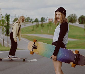 woman with shoulder-length hair holding a rainbow skateboard. another woman skates near her.