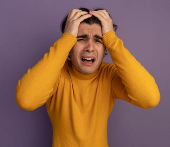 Young man wearing a yellow jumper. He has a pained expression, and both of his hands are clutching his hair in despair. He is standing before a plain purple background.