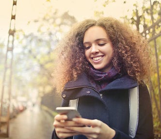 A young, attractive woman with curly brown hair smiling broadly as she looks at her phone.