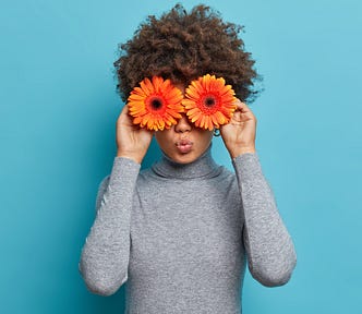 Young woman is holding two large, round, orange gerberas in front of her eyes.. Her lips are rounded. She is wearing grey against a blue background.