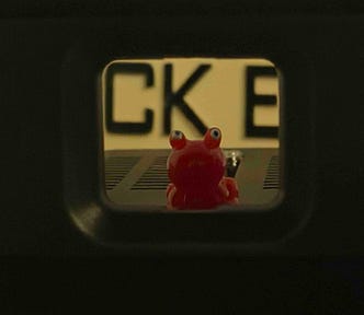 A small red toy frog with large round eyes sits centered inside a narrow rectangular opening, with blurred black letters in the background. The framing creates a tunnel-like view that draws attention to the bright, playful figure.