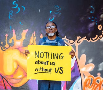 A Deaf Black man with a septum piercing and a black and white beard, wearing denim overalls and glasses, holds a hand-lettered sign that reads ‘NOTHING about us without US.’ A vibrant mural of a Black woman serves as the background.