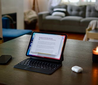 IPad Pro in magic keyboard on dining room table next to AirPods, iPhone, and lit candle.