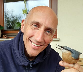 The author, József Manhertz, looks into the camera with a smile. He is bald and has blue eyes. A Eurasian nuthatch sits on the index finger of his left palm.