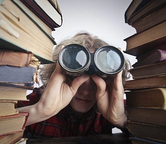 Boy looking through old binoculars sitting between stacks of books.