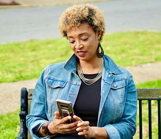 A woman in a denim jacket sits on a park bench and looks at her phone