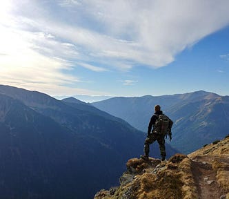 A man with a backpack stands on a trail overlooking remote mountains.