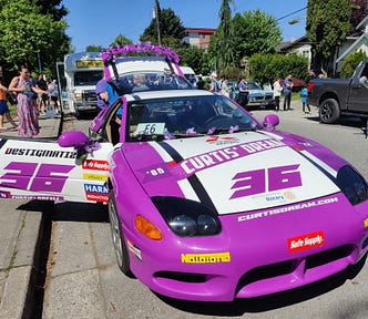 A purple and white sports car parked on the side of a road with people in the background.