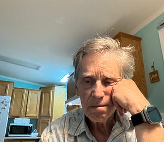 An older man with gray hair sits at a kitchen table, resting his face on his hand and appearing deep in thought or perhaps dejected. He is wearing a plaid shirt and a smartwatch. Behind him are wooden cabinets, a microwave, and a refrigerator with butterfly magnets. The lighting is bright, and the overall mood seems contemplative or sad.