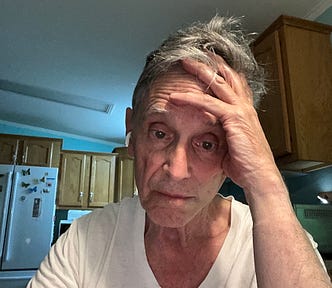 An older man wearing a white T-shirt and wireless earbuds sits in a kitchen, resting his head on one hand and looking slightly weary or contemplative.