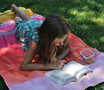 Woman lying on a towel outside on a grassy lawn, reading a book