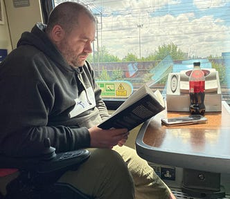 Scott sat reading on a train. On the table in front is a bottle of drink and his phone