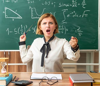 Angry young female teacher sits at table with school tools points at blackboard showing you gesture in classroom
