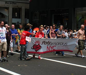 A group of people are marching down a street carrying a sign that says “Polyamorous NYC”. One man on the far left has a t-shirt that says “Date Me and My Husband”. Some other people in the background appear to be queer-coded. One is wearing a tie dye t-shirt. Others are dressed more conservatively.