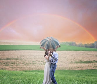 Couple kissing while holding an umbrella in a field with a rainbow in the background.