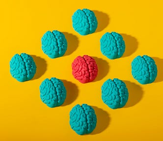 3D illustration of nine human brains: eight in blue and one in bright pink, set against a yellow background. The blue brains are arranged in a diamond shape, with the pink brain standing out at the center.