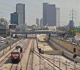 View looking towards buildings in the background, a train is approaching on railway tracks in the mdidle of the photo with highway traffic on either side
