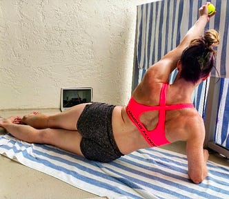 The author lies on her side in a bright pink sports bra and shorts, performing a single arm shoulder CAR with a tennis ball squeezed in her hand