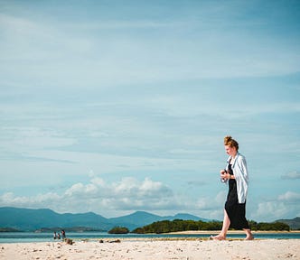 A woman in a black dress with red hair stacked in a bun walking on a sandy short with hills and sky in the background