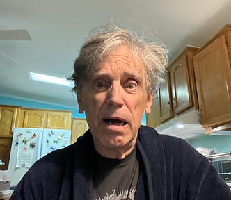 A surprised-looking man with disheveled gray hair wearing a dark robe and a T-shirt, standing in a kitchen. The background shows wooden cabinets, a refrigerator with colorful magnets, and a ceiling fan above. The expression on his face suggests shock or confusion.