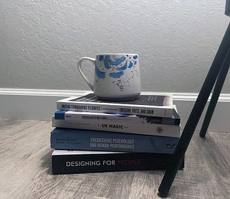 A blue and white coffee mug sits atop a stack of 5 books, on the floor, against a wall. Next to them are the legs of a lamp.