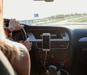 The picture shows a woman driving a car onto a stretch of freeway. The dashboard and accessories are visible. Her cell phone is plugged into a USB port. The article is about the author’s parents’ strategies for approaching traffic tickets. | Photo courtesy: Peter Fazekas, Pexels