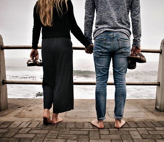 rear view of couple standing on brick sidewalk holding hands carrying shoes looking out to the ocean