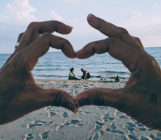 Photo of two hands making a heart in front of the camera, with the beach and water in the background. In the middle of the heart is a couple, sitting in the sand, close to each other.