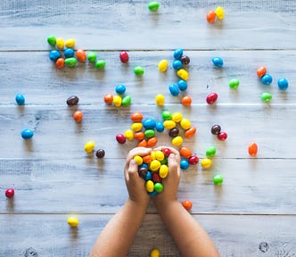 Child’s hands holding jelly beans. Quite a few jelly beans spread across a light colored wooden surface.