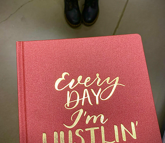 View of a notebook that says “Every DAY I’m Hustlin’” with view of person wearing Doc Martens in the background.