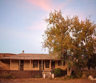 A picture of a small abandoned house in the mining town of Cananea, Mexico at sunset. The sky is blue with tints of rose, it’s about to get dark. The house has an old crooked roof and is boarded up. In front of it a big tree is doused with the last light of the golden hour. The ground around is dry and light brown.