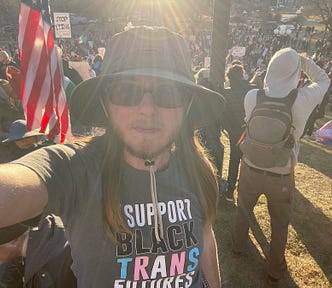 The author is standing in front of a large crowd of people protesting. He’s wearing a hat and sunglasses. He has long brown hair and a beard. His t-shirt says “I support Black trans futures.” There’s an American flag behind him on the left and several protest signs. One sign says, “stop lying”. A person in the background is wearing a trans flag on the right. Another person in the background is waving a Mexican flag.