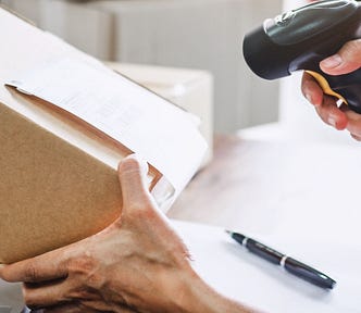 Image of a hand holding a scanner, scanning a shipping label on a box being prepared for shipping.