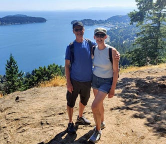A man and woman standing on a rocky viewpoint in summer with a backdrop of ocean and trees.