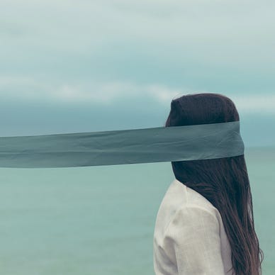 person facing the right with a wide teal ribbon covering their face and trailing far behind them out of the image, person’s head is covered in long, straight dark hair falling onto a white shirt, opaque sea or lake and cloudy sky in the background