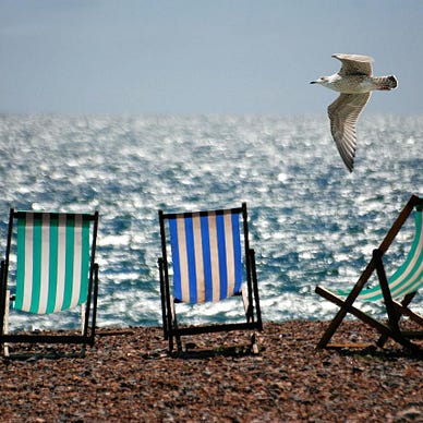 Three-green and blue stripy beach chairs on a pebbled beach with glittering waves in the background and a seagull flying by.