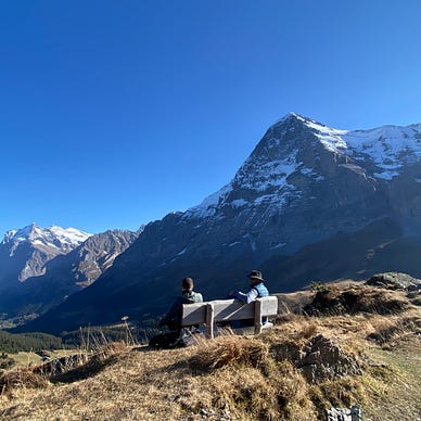 Two people sit on a bench with large mountains in the background and a valley below.