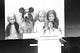 The author stands at a podium with microphone, presenting a slide that says, “What if we’re all just impostors?” The slide features an image of three American Girl dolls in a row alongside a wiener dog in Mickey Mouse ears.