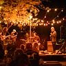 A four person band plays outdoors under a canopy of string lights at night.